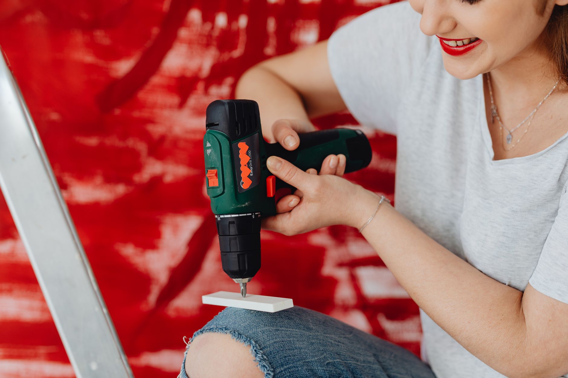 A smiling woman using a cordless drill, engaged in a home renovation project with a red background.